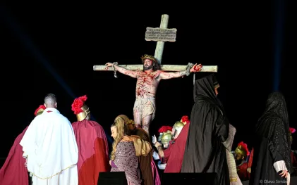 Um ator interpreta a cena do espetáculo A Paixão de Cristo em que Jesus é crucificado. Ele está pregado em uma cruz de madeira e seu corpo está coberto por arranhões com sangue. Ele veste apenas um pano claro que cobre a parte de baixo do seu corpo e usa uma coroa de espinhos. Em cena, ao redor da cruz, também estão atores vestidos como soldados romanos e mulheres vestidas com mantos que cobrem seus cabelos.