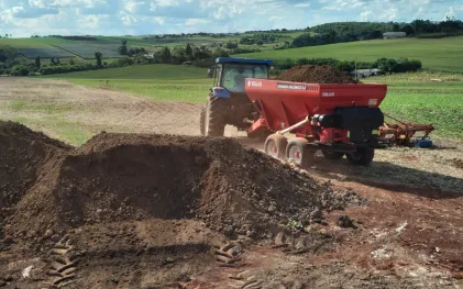 Trator com distribuidor de adubo vermelho sendo abastecido com lodo de esgoto higienizado em uma área agrícola.
