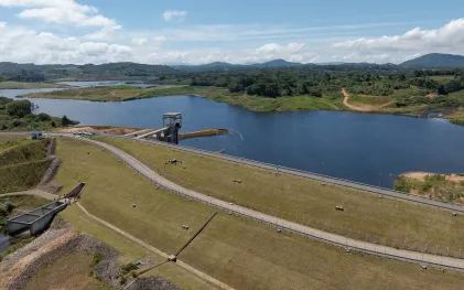 Vista aérea panorâmica da Barragem de Miringuava em um dia ensolarado com nuvens esparsas. À direita, estende-se um grande reservatório de água azul-escura cercado por vegetação nativa, matas e colinas ao fundo. No centro da imagem, o talude de terra da barragem é coberto por grama verde, cortado por vias pavimentadas onde é possível ver pequenos veículos e estruturas técnicas, incluindo uma torre de captação de concreto que avança sobre a água