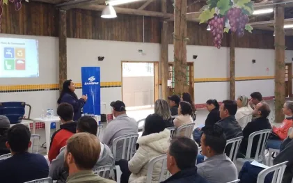 Palestrante da Sanepar realiza palestra sobre eixos do saneamento em espaço comunitário decorado com cachos de uva artesanais, para plateia atenta de moradores.