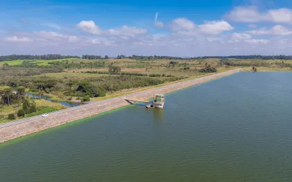 Vista ampla da Barragem do Iraí mostrando o espelho d'água calmo do reservatório, delimitado por uma longa extensão de talude gramado sob o céu com nuvens.