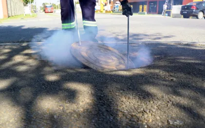 Funcionário da Sanepar utiliza haste metálica para abrir tampa de bueiro de onde sai fumaça densa e esbranquiçada durante teste na rede de esgoto