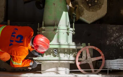 Profissional com uniforme laranja e capacete vermelho realiza a manutenção preventiva ou troca de parafusos em uma tubulação hidráulica verde de grande porte com válvula de roda.