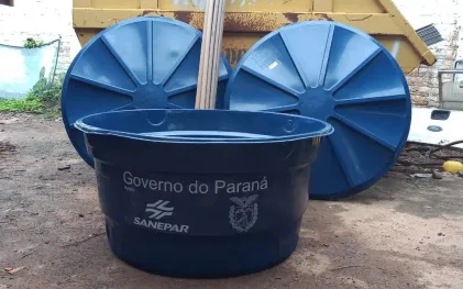 Caixa-d'água azul no chão com as logos do Governo do Paraná e da Sanepar estampadas na frente. Ao fundo, duas tampas azuis estão apoiadas verticalmente contra uma caçamba amarela.