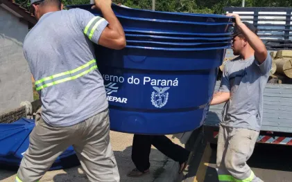 Dois trabalhadores com uniformes cinza carregam uma pilha de caixas d'água azuis. Na lateral da caixa superior, estão estampados o logotipo da Sanepar e o brasão do Governo do Paraná.