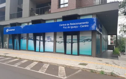 Fachada da Central de Relacionamento da Sanepar em Foz do Iguaçu (Centro), localizada no térreo de um prédio moderno. A fachada possui uma grande faixa azul no topo com a logomarca da Sanepar e os dizeres "Central de Relacionamento Foz do Iguaçu - Centro". As vitrines de vidro exibem o texto "A vida com a Sanepar é melhor" sobre uma ilustração de ondas de água clara