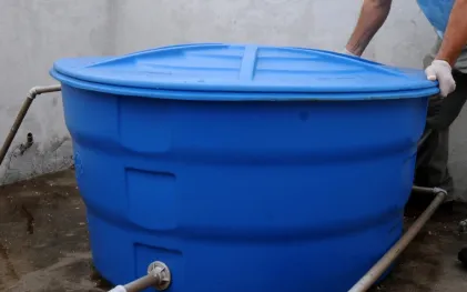 Caixa d'água de plástico azul instalada sobre piso de concreto, com tubulações conectadas à base e uma pessoa de luvas brancas ajustando a tampa.