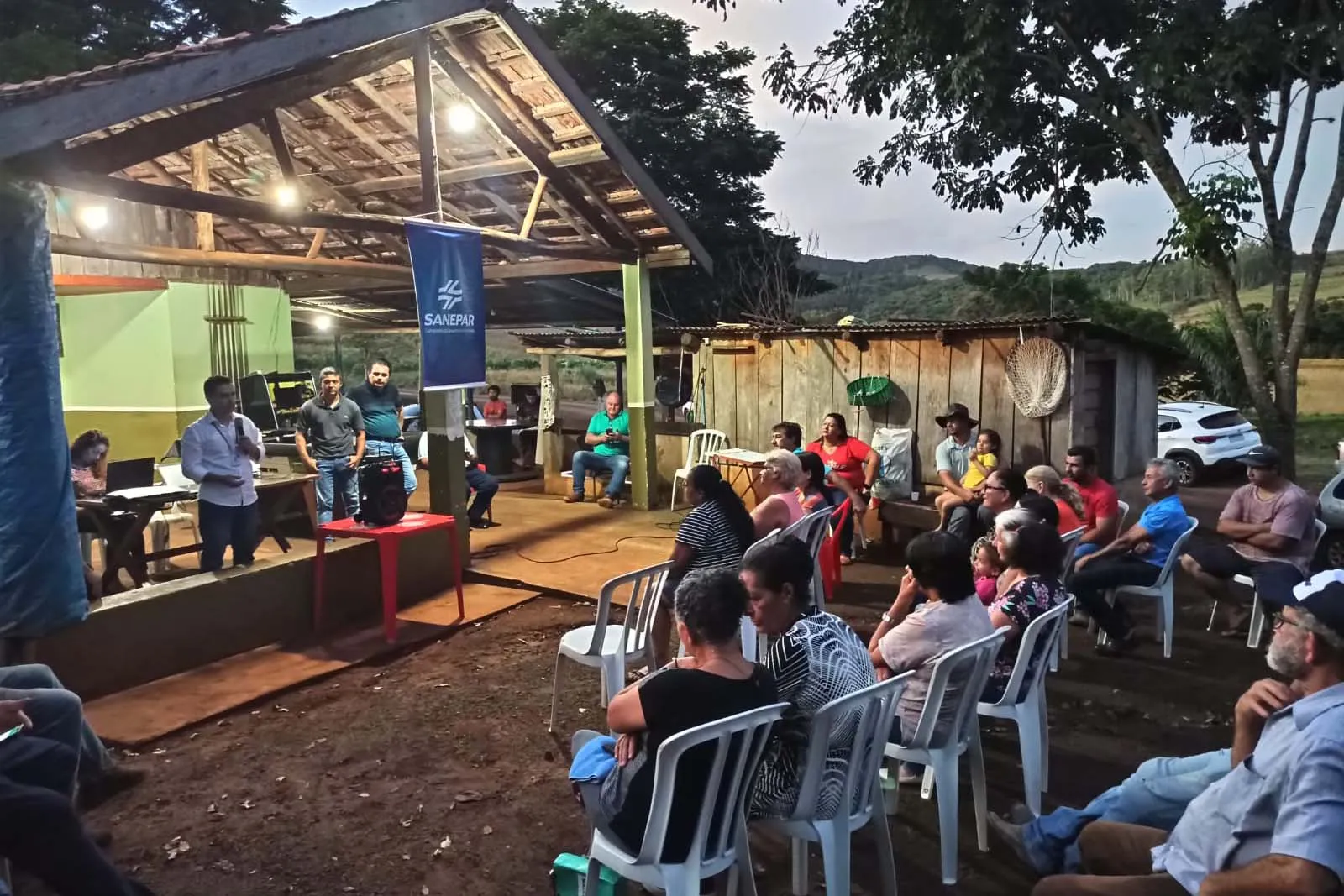 Comunidade de pessoas reunidas em um pequeno galpão e ao ar livre, ouvindo uma palestra.